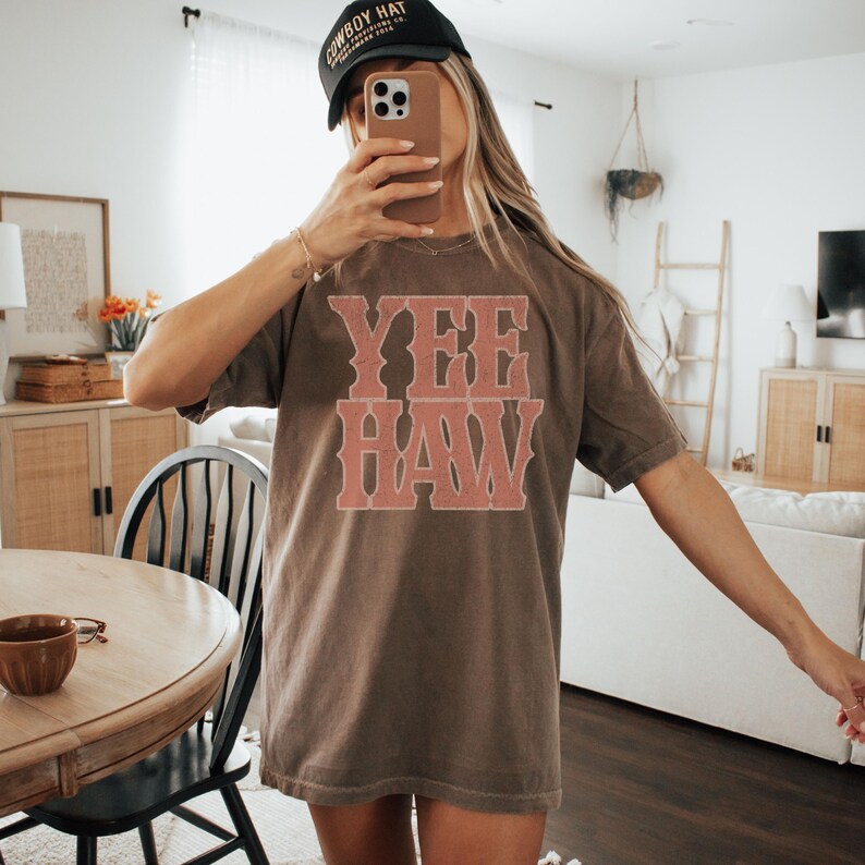 Pu&ograve; includere: Una maglietta marrone con la scritta "YEE HAW" in un carattere color corallo in stile vintage. La maglietta &egrave; di vestibilit&agrave; ampia. Viene indossato un berretto da baseball nero con testo bianco.