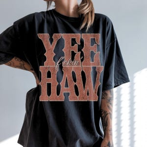 May include: Black t-shirt with the text "YEE f*ckin' HAW" in a distressed, western-style font. The text is in a reddish-brown color, contrasting against the dark fabric. The shirt has a vintage, worn look.