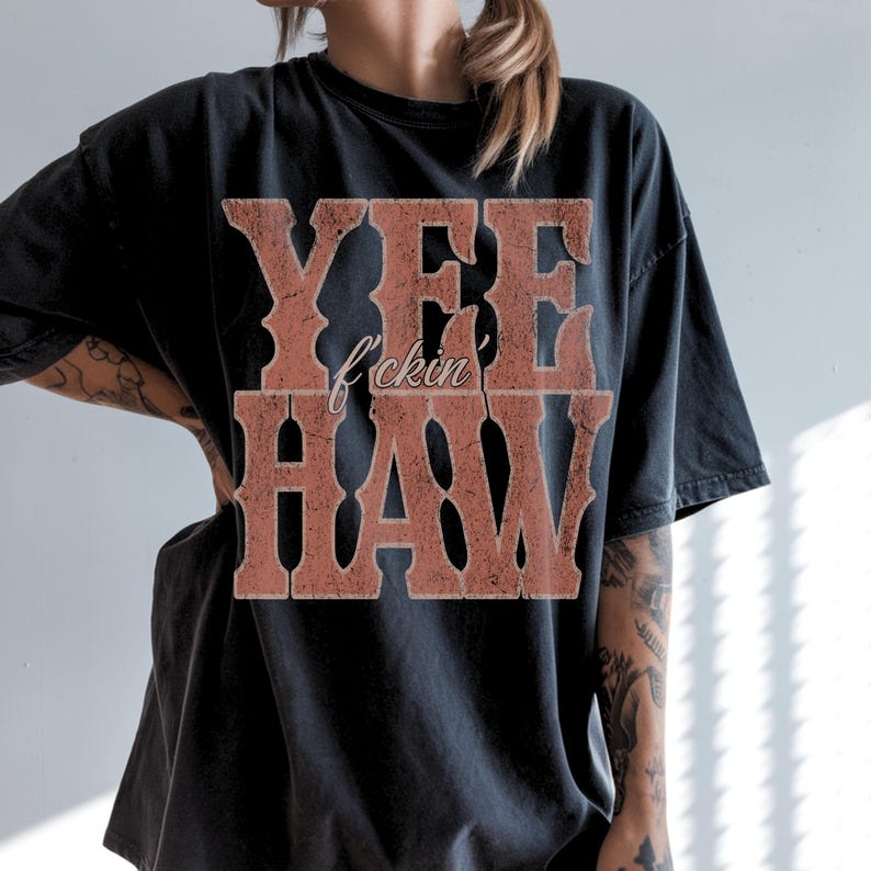 May include: Black t-shirt with the text "YEE f*ckin' HAW" in a distressed, western-style font. The text is in a reddish-brown color, contrasting against the dark fabric. The shirt has a vintage, worn look.