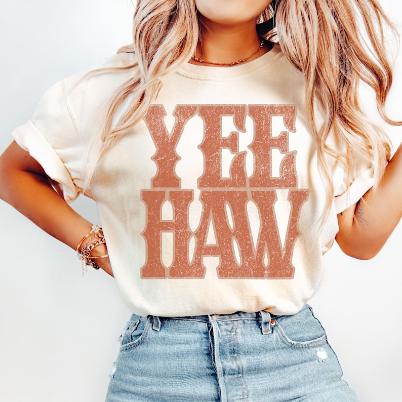 Pu&ograve; includere: T-shirt color crema con la scritta "YEE HAW" in un carattere in stile western color ruggine. La maglietta &egrave; abbinata a jeans blu. L'estetica generale suggerisce un tema country o western.