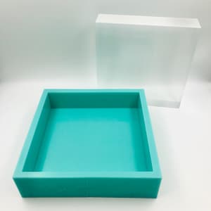 Può includere: Uno stampo in silicone turchese per la colata di resina, con un coperchio in scatola acrilica trasparente. Lo stampo è quadrato e ha un bordo rialzato.