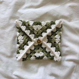 Puede incluir: Bolsa de crochet hecha a mano con un patrón de cuadrados de la abuela. La bolsa presenta una mezcla de cuadrados verdes y blancos, con un borde blanco y un cierre de botón. El artículo se utiliza probablemente para transportar objetos pequeños.