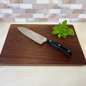 Pode incluir: Uma tábua de corte de madeira castanha escura com uma faca de chef de aço inoxidável e um raminho de manjericão fresco. A faca tem um cabo preto com rebites prateados. A tábua é retangular.