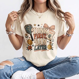 May include: A cream-colored short-sleeve t-shirt featuring a graphic print with the text DINK FIRST BAM LATER. The design includes a pickleball paddle, a pickleball, a mahjong tile with the character for fa, flowers, and a striped bow.