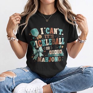 May include: A black t-shirt featuring a graphic design with the text I CAN'T IT'S PICKLEBALL OR MAHJONG. The design includes a pickleball paddle, a ball, a blue plaid bow, and a set of mahjong tiles, all rendered in a vintage-inspired color palette.