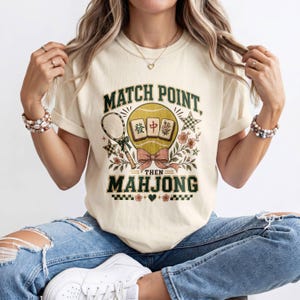 Pode incluir: Uma t-shirt de cor creme com uma estampa gráfica que inclui o texto MATCH POINT, THEN MAHJONG. O design apresenta uma raquete de ténis, uma bola de ténis amarela, peças de mahjong, um laço cor-de-rosa, flores e um pequeno coração verde.