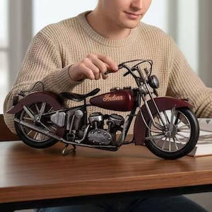 May include: A maroon and silver Indian motorcycle model sits on a wooden table. The model has detailed features, including a seat, wheels, and engine components. The word "Indian" is visible on the gas tank. The motorcycle is a decorative item.