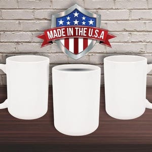 May include: Three white ceramic mugs and a white cylindrical cup are displayed on a dark brown surface against a brick wall. A shield-shaped emblem with the text "MADE IN THE U.S.A." is visible above the cups.