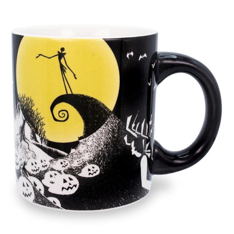May include: Black ceramic mug featuring a design from The Nightmare Before Christmas. The mug has a black handle and a white interior. The design includes Jack Skellington standing on a hill in front of a yellow moon, with bats and pumpkins.