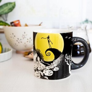 May include: A ceramic mug featuring a black and white illustration of a scene with a yellow moon, a silhouetted figure, and jack-o'-lanterns. The mug has a black handle and a white interior. The mug is on a white surface.