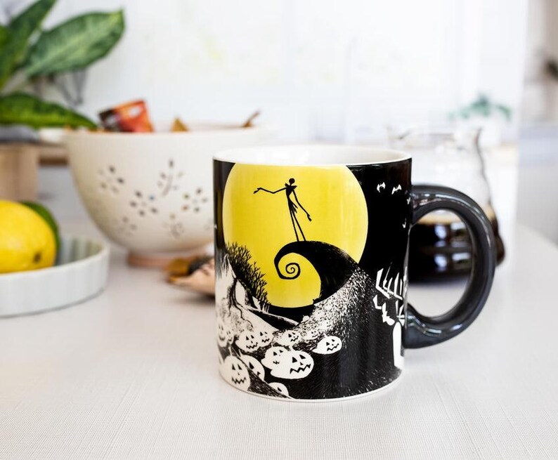May include: A ceramic mug featuring a black and white illustration of a scene with a yellow moon, a silhouetted figure, and jack-o'-lanterns. The mug has a black handle and a white interior. The mug is on a white surface.