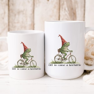 May include: Two white ceramic mugs with a whimsical illustration of a green frog riding a bicycle and wearing a red hat. The text "Off to cause a kerfuffle" is printed below the image. One mug is larger than the other.