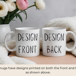 May include: Two white ceramic mugs with black text that reads "DESIGN FRONT" and "DESIGN BACK". The mugs are on a light pink surface, with a white vase of flowers in the background. The flowers are white and pink.
