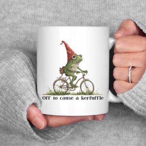 May include: White ceramic mug featuring a whimsical illustration of a green frog riding a bicycle, wearing a red gnome hat. The text "Off to cause a kerfuffle" is printed below the image. The mug is held by a person.