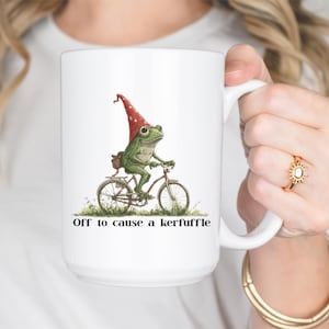 May include: White ceramic mug featuring a whimsical illustration of a green frog riding a bicycle and wearing a red gnome hat. The mug has the text "Off to cause a kerfuffle" printed on it.