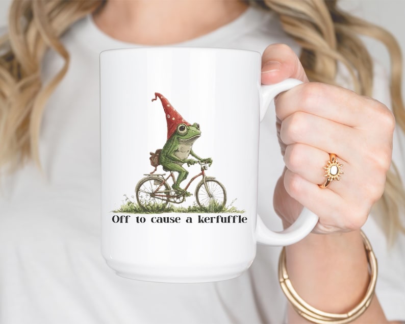 May include: White ceramic mug featuring a whimsical illustration of a green frog riding a bicycle and wearing a red gnome hat. The mug has the text "Off to cause a kerfuffle" printed on it.