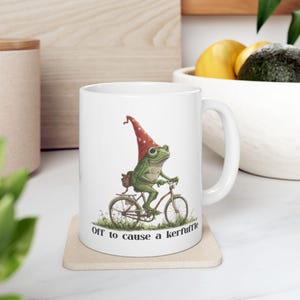 May include: White ceramic mug featuring a whimsical illustration of a green frog wearing a red gnome hat and riding a bicycle. The text "Off to cause a kerfuffle" is printed below the image. The mug has a curved handle.