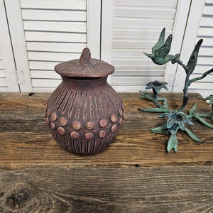 May include: A maroon ceramic jar with a lid, featuring vertical grooves and circular accents. The jar sits on a wooden surface, alongside a bronze bird sculpture. The background includes white shutters.