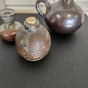 May include: Three ceramic vessels in shades of brown, green, and blue. The central piece is a round bottle with a cork stopper. The other two are small vases, one with a handle. The pottery has a rustic, handmade appearance.