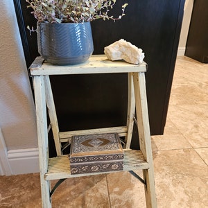 May include: A white painted wooden step ladder with a decorative box on the top step. The ladder is sitting on a tiled floor. A small potted plant is on the top step of the ladder.