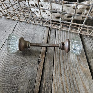 May include: A pair of antique glass doorknobs with a metal rod connecting them. The doorknobs are clear glass with a faceted design and the rod is rusty.