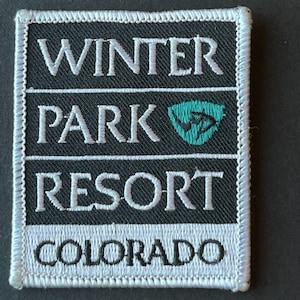 May include: A rectangular patch with the words "WINTER PARK RESORT COLORADO" embroidered in white on a black background. A teal-colored emblem is featured between "PARK" and "RESORT". The patch has a white border.