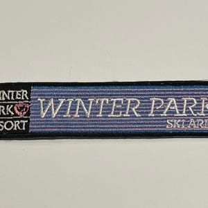 May include: Embroidered patch with the words "Winter Park Resort" and "Ski Area" in white lettering on a blue and pink striped background. The patch has a black border.