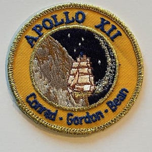 May include: Embroidered patch with a gold border and a yellow background. The patch features the words "APOLLO XII" in blue, along with the names "Conrad - Gordon - Bean." The central design depicts a ship on the moon.