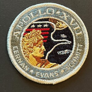 May include: Embroidered patch with the text "APOLLO XVII" and the names "GERNAN * EVANS * SCHMITT". The patch features a profile of a person, a stylized American flag, and a depiction of space. The patch has a gray border.