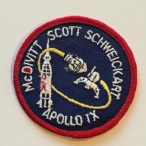 May include: A circular patch with a navy blue background and a red border. The patch features a rocket, astronaut, and the text "McDivitt Scott Schweickart Apollo IX". The design is embroidered with white and yellow thread.