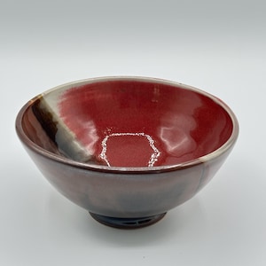 Pode incluir: Uma tigela de cerâmica feita à mão com acabamento brilhante. O interior é vermelho vibrante, passando para um castanho mais escuro na base. A borda apresenta um gradiente branco e castanho. A tigela tem cerca de 15 cm de diâmetro.