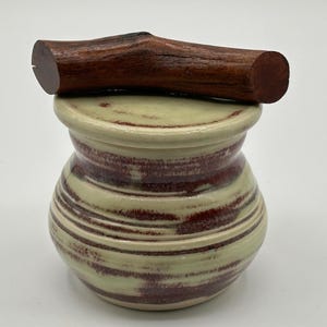 Peut inclure: Un pot en céramique artisanal avec un couvercle en branche de bois brun foncé. Le pot présente un motif tourbillonnant vert clair et marron. Le pot est un objet décoratif.