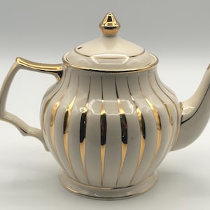 Puede incluir: Tetera crema con rayas verticales y detalles dorados. La tetera tiene una tapa a juego con un adorno dorado y un pico curvo. El asa y el pico también están adornados con oro. Un diseño clásico para servir té.