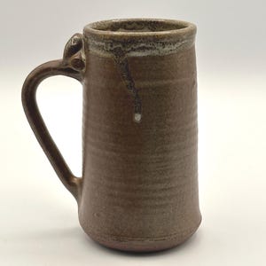 May include: A handcrafted brown ceramic mug with a large handle. The mug has a cylindrical shape with a slightly flared base. The rim has a lighter brown color with a drip design. The mug is approximately 6 inches tall.