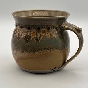 Pode incluir: Uma caneca de cerâmica artesanal com um design único. A caneca apresenta um esquema de cores castanho e bege com uma superfície texturizada. Possui uma alça e orifícios decorativos ao redor da borda. Adequada para café, chá ou outras bebidas.