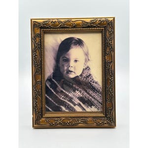 May include: A rectangular picture frame with a metallic gold finish and an ornate, embossed floral vine pattern along the border. The frame contains a black and white photograph of a child wrapped in a textured, striped knit blanket or shawl.