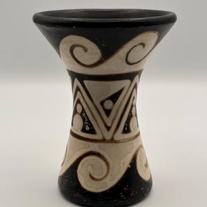 Puede incluir: Un jarrón decorativo de cerámica con una parte superior acampanada y una base estrecha. El jarrón presenta un borde y una base negros, con un cuerpo de color crema adornado con patrones de remolinos y geométricos marrones y crema. El jarrón mide unos 15 cm de alto.