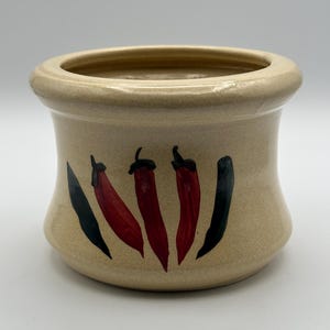 Pode incluir: Um recipiente de cerâmica bege com uma borda larga e um corpo curvo. O recipiente é decorado com três pimentas vermelhas e duas pimentas pretas. As pimentas são pintadas com caules pretos e folhas verdes.