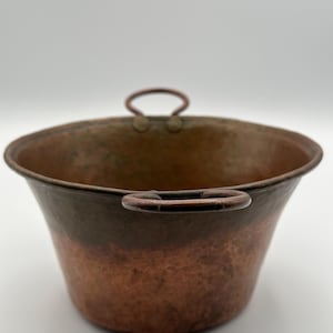 May include: A copper bucket with two small handles and a rounded rim. The bucket has a hammered texture and a gradient of colours, from a dark brown at the top to a bright copper at the bottom. The bucket is empty and sits on a white surface.