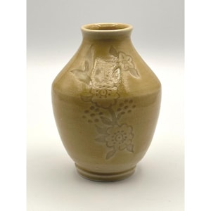 Puede incluir: Un pequeño jarrón de cerámica de forma bulbosa con un esmalte brillante de color miel. La superficie presenta un sutil patrón floral en relieve que representa un tallo central con flores y pequeñas bayas, extendiéndose desde la base.