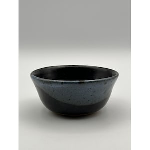 Pode incluir: Uma tigela de cerâmica artesanal com esmalte azul escuro e preto. A tigela tem formato arredondado e textura salpicada, perfeita para servir sopas, saladas ou lanches. O interior é preto profundo, contrastando com os tons azuis do exterior.