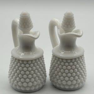 Puede incluir: Dos botellas de vidrio blanco con textura de clavo, con asa y tapón decorativo. Las botellas tienen una base bulbosa y un cuello delgado. Los tapones son cónicos. Los aceiteros se muestran sobre un fondo blanco.