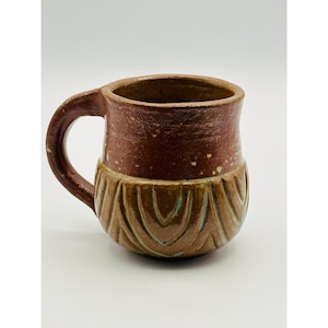 Pode incluir: Caneca de cerâmica artesanal em tons de castanho e bege. A parte superior é castanho escuro com salpicos brancos, a secção inferior apresenta um design texturizado e esculpido em bege e turquesa. Com uma pega arredondada.
