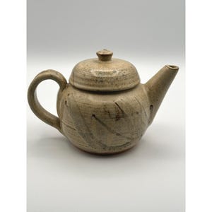 Pode incluir: Bule de cerâmica bege com tampa e alça. O bule tem um bico e é decorado com riscas castanhas escuras. O bule é provavelmente feito à mão e adequado para preparar chá.