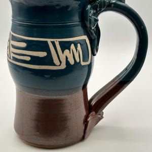 May include: A handcrafted ceramic mug with a teal glaze on the upper portion and a brown base. The mug features a unique handle design and a cream-colored geometric pattern around the middle. The mug is approximately 5 inches tall.