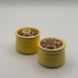 Pode incluir: Dois pequenos potes de cerâmica amarelos com tampas. As tampas são marrons com um desenho floral em amarelo e marrom. Os potes são cilíndricos com uma base ligeiramente alargada. O fundo é branco liso.