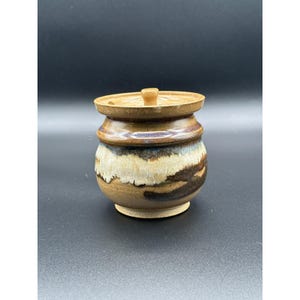 May include: A small, round ceramic jar with a matching lid featuring a central knob. The vessel displays a multi-toned glaze with earthy brown, cream, and dark blue hues, characterized by a horizontal drip pattern around the center body.