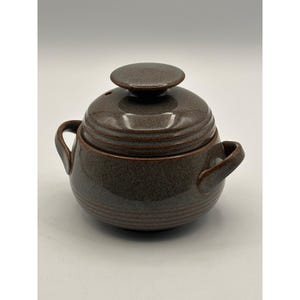 Op de afbeelding: Een bruine keramische braadpan met deksel en twee handvatten aan de zijkant. Het deksel heeft een kleine knop en een ventilatiegat. De schaal heeft een ronde vorm met horizontale ribbels.