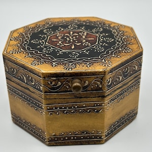 Puede incluir: Caja octogonal de madera con tapa con bisagras. La caja está pintada de dorado con intrincados diseños florales en blanco y negro en la tapa y los lados. Un pequeño pomo redondo sirve de cierre.
