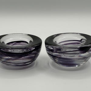 Puede incluir: Dos candelabros de vidrio transparente con un diseño de remolino en tonos morados y grises. Los candelabros tienen forma redonda con una abertura ancha y un borde grueso. El vidrio tiene un acabado brillante.